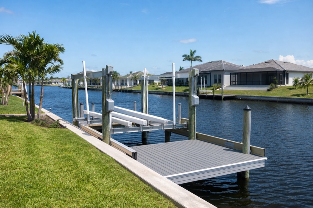 An empty aluminum boat lift with white guide poles beside a grey composite dock platform, with a concrete seawall running along the lawn behind it