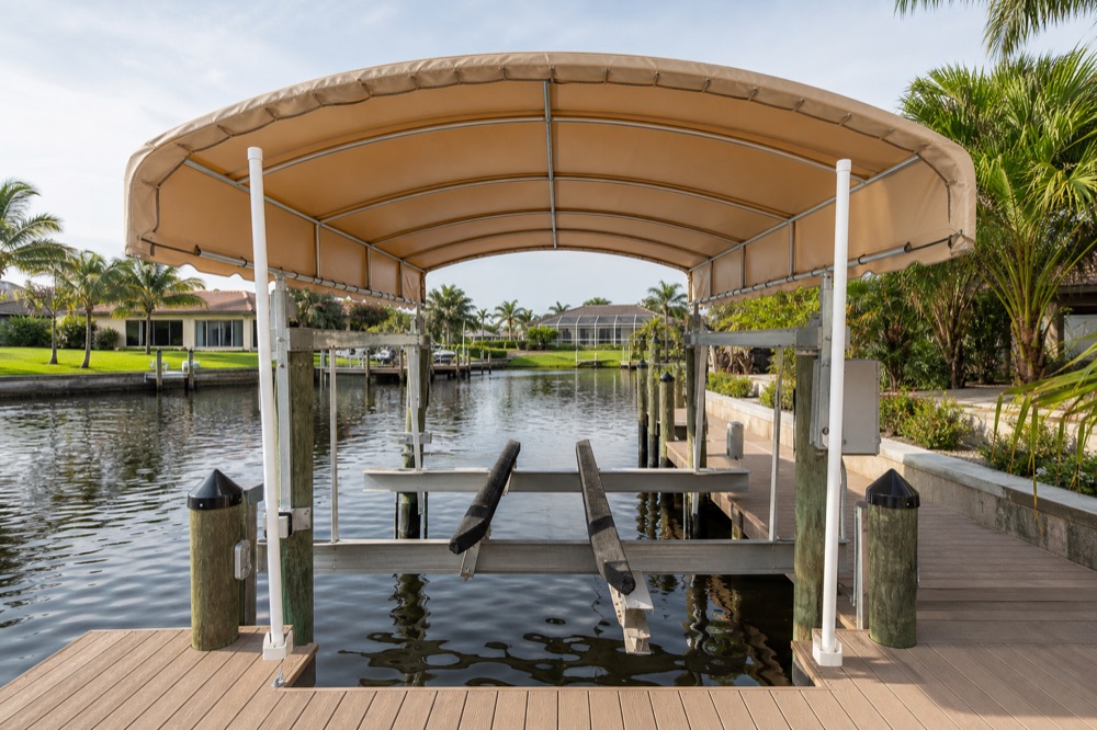 An empty covered boat lift with a tan canvas canopy on an aluminum frame, seen from a composite dock deck across a wide calm canal