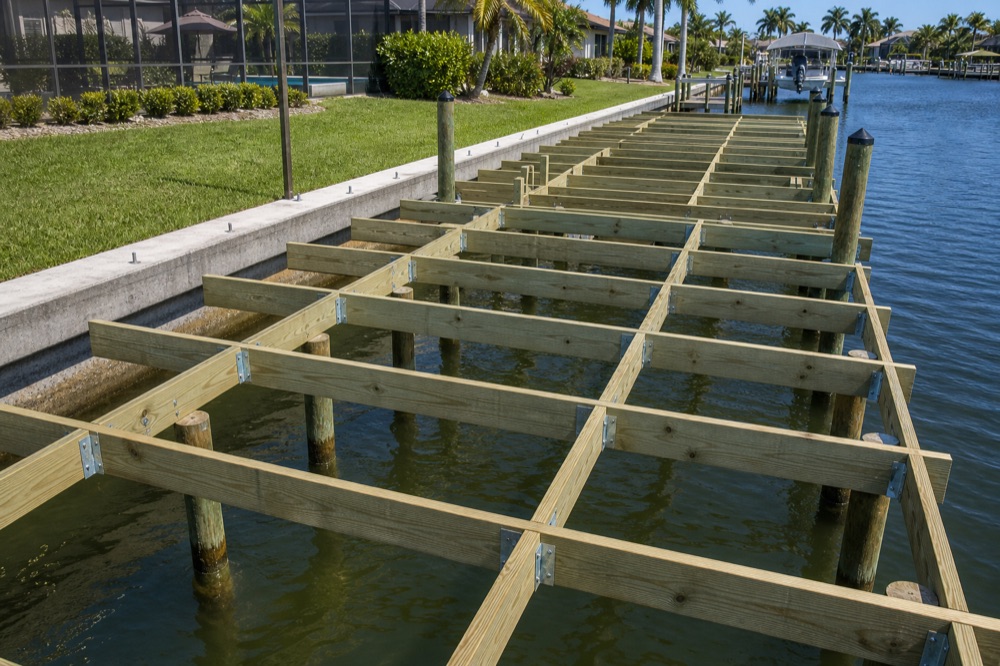 The open frame of a dock under construction over a Southwest Florida canal, stringers and joists bracketed to pilings alongside a finished concrete seawall, before any decking is laid